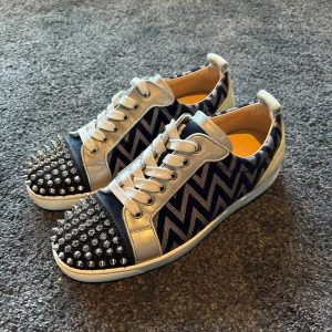 Christian Louboutin sneakers med nitar - Unika sneakers från Christian Louboutin med silverfärgade detaljer, svart och vit zigzag-mönster på sidorna och coola nitar på tån. Skorna har klassiska röda sulor och snörning. Materialmix av skinn och textil ger en lyxig känsla. Skick: 8,5/10
