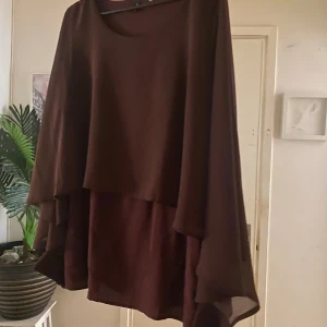 Vinröd omlottklänning med volang - Flödande vinröd klänning med lager på lager-effekt och vida, transparenta ärmar. Klänningen har en snygg omlottkänsla och volangdetaljer som ger extra rörelse. Perfekt för dig som gillar en luftig och elegant stil.