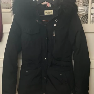 Svart vinterjacka Rocco Barocco med fuskpäls - Svart vinterjacka från Rocco Barocco med fluffig fuskpäls på huvan, dragkedja och knappar framtill samt flera fickor. Jackan har en figurnära passform och är perfekt för kalla dagar. Snygg och stilren design som håller dig varm hela vintern.