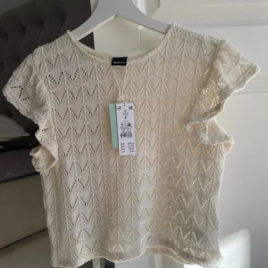 Vit spetsblus från Gina Tricot - Supersöt vit blus från Gina Tricot med spets och korta volangärmar. Blusen har ett genombrutet mönster och rund halsringning. Perfekt för dig som gillar romantisk och feminin stil.