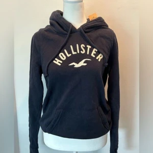 Mörkblå hoodie från Hollister - Säljer en mörkblå hoodie från Hollister med vit logotyp och fågeltryck på bröstet. Tröjan har huva med snörning och en klassisk känguruficka framtill. Perfekt för chill dagar och enkel att matcha med jeans eller leggings. Är strl xs men skulle säga att den passar s oxå 