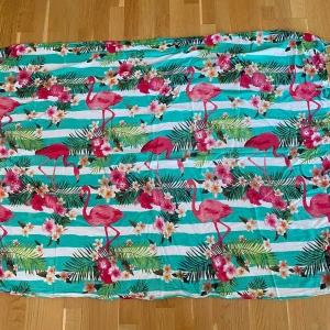Turkos sarong med flamingos och blommor - Säljer en färgstark sarong med turkosa och vita ränder, rosa flamingos och tropiska blommor. Är gjord i ett lätt bomullsmaterial, perfekt för varma dagar. Mönstret är riktigt somrigt och sticker ut med sina tropiska vibes. Mått Längd: ca 150 cm Bredd: ca 107 cm  Kommer från ett djur och rökfritt hem. 