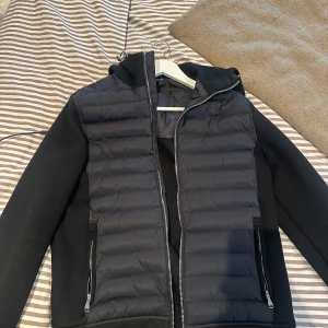 Svart pufferjacka Alessandro Zavetti - Svart pufferjacka från Alessandro Zavetti med huva och dragkedja. Jackan har quiltad framsida, släta ärmar och två fickor med dragkedja. Snygg och modern look, perfekt för dig som vill ha en stilren jacka med edge.