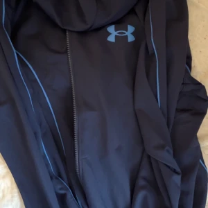 Svart hoodie från Under Armour - Snygg svart hoodie från Under Armour med dragkedja och blå logga på bröstet. Hoodien har tunna blå detaljer längs sömmarna och är tillverkad i ett sportigt, mjukt material. Perfekt för dig som gillar en clean och sportig stil.