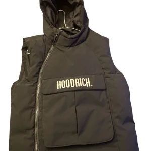 Svart dunväst från Hoodrich XL - Svart dunväst från Hoodrich med huva och stor ficka framtill med HOODRICH-logga. Västen har sned dragkedja och stilren design, perfekt för att ge din outfit en urban touch. Materialet är polyester och västen är quiltad för extra värme.
