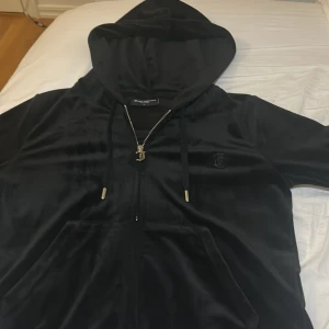 Svart velour hoodie från Juicy Couture - Svart hoodie i mjuk velour från Juicy Couture med dragkedja och huva. Tröjan har snörning med gulddetaljer, fickor framtill och ett broderat JC-logo på bröstet. Perfekt för en chill och trendig look. Använt 3 gånger, kommer ej till användning längre. Storlek L men mer som M