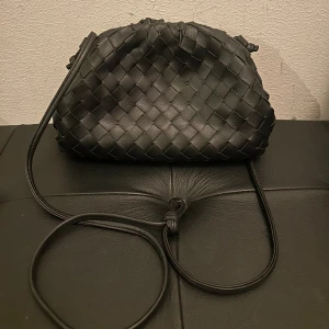 Svart axelväska från Pitaya i skinn - Snygg svart axelväska från Pitaya med flätad design och lång smal axelrem. Väskan är tillverkad i skinn och har en mjuk, rundad form med dragsko upptill. Perfekt för dig som gillar stilrena detaljer och vill ha en väska som sticker ut.