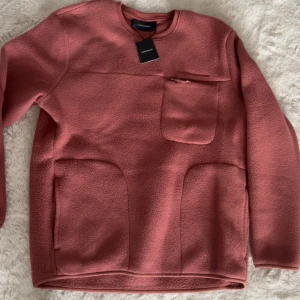  Peak Performance fleece-tröja i gammelrosa/rost  med fickor - Mysig rosa fleece-tröja från Weekday med rund hals, lång ärm och praktiska fickor framtill. Bröstficka med dragkedja ger extra stil och funktion. Perfekt för kyliga dagar och enkel att matcha med jeans eller leggings.