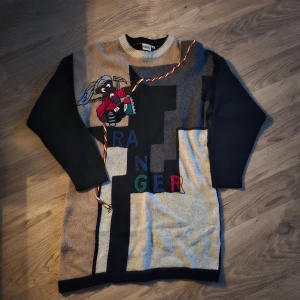 Färgglad stickad tröja från Creation Camilla - Unik stickad tröja från Creation Camilla med coolt motiv av en figur med rep och motorsåg samt texten 'RANGER' i olika färger. Tröjan har svart rygg och ärmar, beige och grå framsida med grafiskt mönster. Rund halsringning och långärmad modell. Storlek M