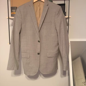 Grå kavaj  - Snygg grå kavaj med två knappar framtill och klassisk krage. Kavajen har två fickor med lock och en bröstficka. Fodrad insida och diskret mönster i tyget. Perfekt för dig som vill ha en stilren och tidlös look.