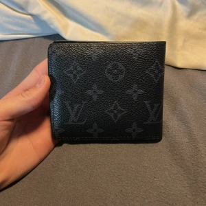 Svart plånbok i skinn - Säljer en svart plånbok från, med klassiskt monogrammönster. Plånboken är tillverkad i skinn och har flera kortfack samt ett fönster för ID eller foto. Perfekt storlek för fickan och riktigt snygg design.