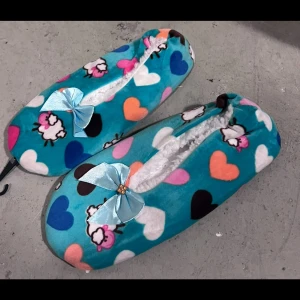 Turkosa ballerinaskor med hjärtan - Mysiga ballerinaskor i turkos med färgglada hjärtan och små får. Insidan är fodrad med vit fleece och ovansidan har en ljusblå rosett. Perfekta att glida runt hemma i, med mjuk och skön känsla. Söt design som sticker ut och gör dig glad varje dag.