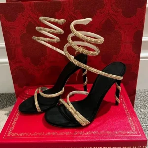 Svarta sandaler med guldspiral från Versace - Unika svarta sandaler från Versace med höga klackar och glittrande guldfärgade spiralband som snor sig upp runt vristen. Skorna har öppen tå och tunna remmar dekorerade med små stenar för extra bling. Perfekta för att sticka ut och ge din outfit en lyxig touch.