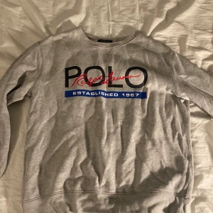 Grå sweatshirt från Polo Ralph Lauren - Snygg grå sweatshirt från Polo Ralph Lauren med stort tryck framtill i svart, rött och blått. Klassisk rund halsringning och långa ärmar. Perfekt för en chill och stilren look.