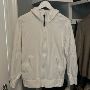 Vit hoodie med goggles från C.P. Company - Vit hoodie från C.P. Company med dragkedja framtill och två fickor med dragkedja. Jackan har huva med inbyggda svarta goggles och en svart detalj på dragkedjan. Tillverkad i mjukt material, perfekt för en cool streetwear-look.