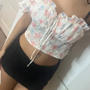 Blommig croppad blus med volang - Säljer en söt croppad blus med blommigt mönster i ljusblått, rosa och vitt. Toppen har volangdetaljer vid axelbanden och runt halsen, samt snörning framtill. Materialet är lätt och luftigt, perfekt för varma dagar. Supersnygg till kjol eller jeans!