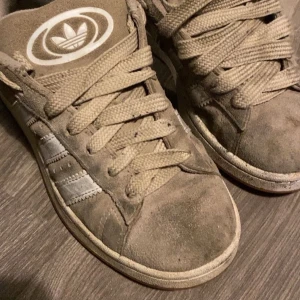 Beige Adidas sneakers med vita ränder - Säljer ett par beige Adidas sneakers med klassiska vita ränder på sidorna och Adidas-logga på plösen. Skorna har snörning och en platt gummisula i orange. Ovandelen är i mocka och har rund tå. Perfekt för dig som gillar retrostil och streetwear.