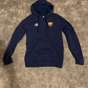 Blå Adidas Djurgården hoodie M - Snygg marinblå hoodie från Adidas med Djurgårdens IF-emblem på bröstet. Tröjan har huva med snörning, stor magficka och långa ärmar med diskret vit detalj längs sidan. Perfekt för dig som vill visa klubbkänsla och hålla dig varm.