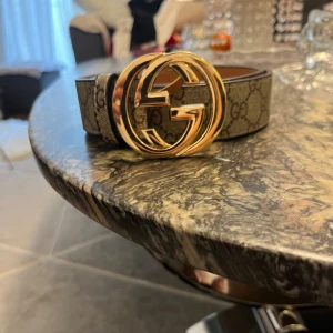  Supreme bälte med guldspänne - Säljer ett klassisk Supreme bälte med stort guldigt -spänne. Bältet är beige med bruna -loggor över hela bandet och har en exklusiv känsla. Perfekt accessoar för att lyfta vilken outfit som helst.