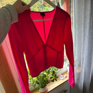 Röd v-ringad blus från Gina Tricot - Säljer en snygg röd blus från Gina Tricot med djup v-ringning och långa ärmar. Blusen har knappar framtill och är gjord i ett lätt, transparent material som ger en cool och trendig look. Perfekt för dig som gillar färg eller ska på pink party 