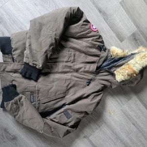 Armygrön dunjacka från Canada Goose - Säljer en Army green dunjacka från Canada Goose med svart ribbstickad mudd vid ärmslut och midja. Jackan har en huva med fuskpäls och dragkedja framtill. Klassisk patch på axeln och flera praktiska fickor. Perfekt för kalla dagar. Muddarna i armsluten behöver bytas. 