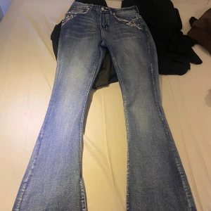 Blå bootcut jeans med döskalle-detaljer - Blå bootcut jeans från SKY med snygga pärl- och nitar på fickorna samt broderade döskallar med vingar på bakfickorna. Jeansen har slitningar och markerade sömmar, samt coola detaljer framtill. Perfekta för dig som vill sticka ut med din stil.