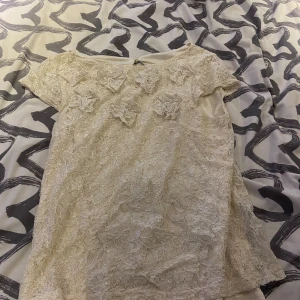Beige spetsblus med blommor - Säljer en beige blus med korta ärmar och vackert spetsmaterial. Framsidan har dekorativa spetsblommor som ger en romantisk vibe. Blusen har rund hals och en liten knapp i nacken. Perfekt för dig som gillar detaljer och mjuka färger.