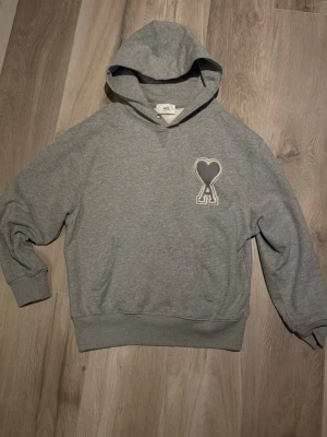 Grå hoodie från Ami Paris - Snygg grå hoodie från Ami Paris med deras ikoniska hjärta-A-logga broderad på bröstet. Klassisk passform med huva och känguruficka. Tillverkad i mjuk bomullsblandning som känns skön mot huden. Perfekt för en chill och stilren look.
