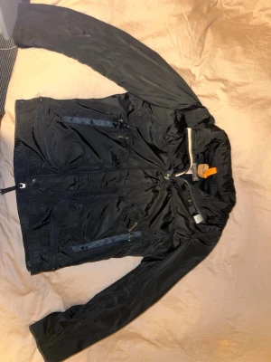 Svart vindjacka från Parajumpers - Snygg svart vindjacka från Parajumpers med dragkedja framtill och flera praktiska fickor. Jackan har meshfoder, hög krage och en orange detalj i nacken. Perfekt för blåsiga dagar och enkel att matcha med olika outfits.