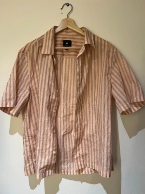 Randig kortärmad skjorta från H&M - Säljer en beige och ljusbrun randig skjorta från H&M. Skjortan har korta ärmar, klassisk krage och knäppning framtill. Tillverkad i tunn bomull, perfekt för varma dagar. Snygg att bära öppen över ett linne eller t-shirt.
