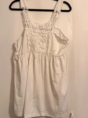 Vit linnetopp med spetsdetaljer - Säljer en söt vit linnetopp med breda axelband och vacker spets framtill. Toppen har en avslappnad passform och är perfekt för varma dagar. Materialet känns mjukt och luftigt, och spetsen ger en romantisk touch.