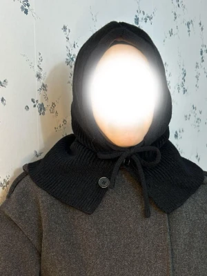 Balaklava  - Säljer en svart stickad huva i mjukt material med bred krage och knytband framtill. Huvan har en knappdetalj vid halsen och täcker både huvud och axlar. Perfekt för dig som vill ha en unik accessoar till höst- och vinterlooken.
