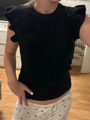 Svart ribbad topp med volangärm - Snygg svart ribbad topp med volangdetaljer på axlarna. Toppen är ärmlös och har en rund halsringning. Jätte skönt material och knappt använd!