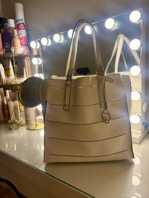 Vit väska från Gerry Webber - Snygg vit väska från Tory Burch/Gerry Webber med perforerade ränder och ett hängande logotyp-hänge. Väskan har två långa handtag och en brun innerpåse med dragkedja. Perfekt för dig som vill ha plats för allt och ändå se stilren ut.