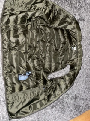 Olivgrön dunväst med dragkedja - Snygg olivgrön dunväst med quiltad design och dragkedja framtill. Västen har två sidofickor med dragkedja och en hög krage. Perfekt att slänga över en hoodie eller tröja för extra värme. Materialet är glansigt och lätt, vilket gör den enkel att bära.