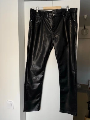 Svarta byxor i skinnimitation - Säljer ett par svarta byxor i skinnimitation med klassisk femficksmodell och raka ben. Byxorna har knapp och dragkedja framtill samt bälteshällor. Snyggt glansigt material som ger en edgy look.