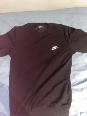 Svart sweatshirt från Nike - Klassisk svart sweatshirt från Nike med rund halsringning och vit Nike-logga broderad på bröstet. Tröjan har ribbade muddar vid ärmslut och nederkant, perfekt för en clean och sportig look. Tillverkad i mjukt bomullsmaterial.