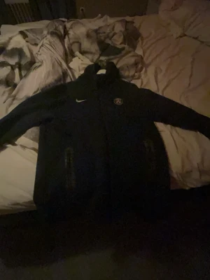 Svart PSG Nike ziptröja - Svart ziptröja från Nike med Paris Saint-Germain-logga på bröstet. Tröjan har hög krage, dragkedja framtill och två fickor med dragkedja. Perfekt för dig som gillar sportig stil och vill representera PSG.