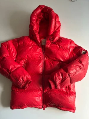 Röd dunjacka från Moncler med huva - Snygg röd dunjacka från Moncler med puffig passform och stor huva. Jackan har dragkedja framtill, ribbade muddar och Moncler-logga på ärmen. Perfekt för kalla dagar när du vill hålla stilen och värmen.