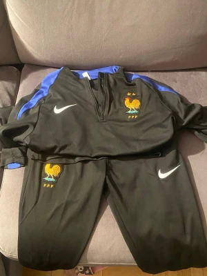 Frankrike Nike träningsset svart/blå - Säljer ett svart träningsset från Nike med Frankrikes landslagslogga (FFF) och två stjärnor. Setet har blå detaljer på axlarna och dragkedja vid halsen. Både tröja och byxor har broderade Nike-loggor och tuppemblem. Perfekt för fotbollsträning.