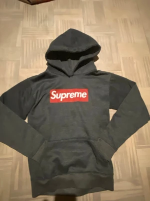 Mörkgrå Supreme hoodie med box logo - Säljer en mörkgrå hoodie från Supreme med den klassiska röda box logon på bröstet. Hoodien har huva och en stor magficka framtill. Perfekt för dig som gillar streetwear och vill sticka ut med en ikonisk look.
