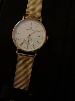 Carl Edmond guldmesh klocka - Stilren klocka från Carl Edmond med vit urtavla och guldfärgat mesharmband i metall. Klockan har minimalistisk design, tunna visare och en liten sekundvisare. Perfekt accessoar för dig som gillar klassisk och modern stil.