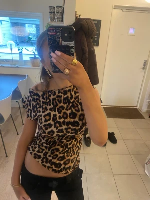 Leopardmönstrad offshoulder topp - Trendig offshoulder topp med leopardmönster i bruna och svarta toner. Figurnära passform och korta ärmar, perfekt för dig som vill sticka ut. Matcha med svarta jeans för en edgy look.