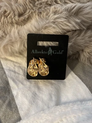 Guldfärgade örhängen Albrekts Guld - Säljer ett par guldfärgade örhängen från Albrekts Guld med vågig, organisk form och glansig finish. Örhängena har en chunky design som ger ett trendigt intryck och är perfekta för att lyfta vilken outfit som helst.