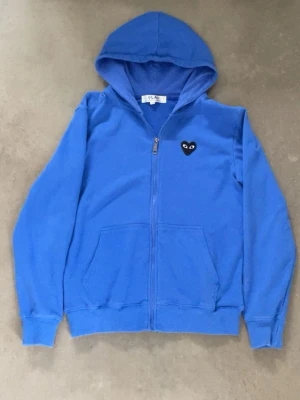 Blå Comme des Garçons PLAY hoodie - Blå hoodie med dragkedja från Comme des Garçons PLAY. Har en svart hjärtpatch med ögon på bröstet och ett stort svart hjärta med ögon på ryggen. Utrustad med huva och kängurufickor. Tillverkad i mjukt bomullsmaterial för skön känsla.