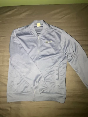 Ljusblå vintage ziptröja från Nike - Snygg ljusblå ziptröja från Nike med diskret logga på bröstet och 'Nike Athletic Club' tryck på ryggen. Tröjan har ribbade muddar, ståkrage och två fickor framtill. Perfekt för en sportig och vintage stil.