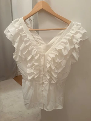 Vit volangblus med v-ringning - Supersöt vit blus med djup v-ringning både fram och bak. Blusen har breda volangdetaljer längs axlarna och är ärmlös. Tillverkad i ett lätt tyg som ger en luftig känsla. Perfekt att styla med jeans eller kjol för en trendig look.