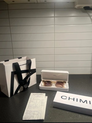 Chimi 04 solglasögon  - Säljer ett par stilrena solglasögon från Chimi med transparent beige båge och bruna tonade glas. Modellen har en fyrkantig form och levereras i originaletui med tillhörande förpackning och kvitto. Perfekt accessoar för en trendig look.