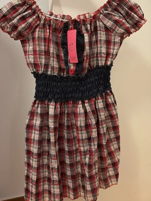 Rutig offshoulder blus - Trendig offshoulder blus i röd, svart och vit rutigt mönster. Blusen har en bred smock i midjan och korta puffärmar. Tillverkad i ett lätt och luftigt tyg, perfekt för en avslappnad och cool stil. Vet inte vilken storlek men troligen passar den Xs-M.