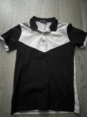 Svart och grå pikétröja från Prada - Snygg pikétröja från Prada i svart och grått med klassisk krage och knappslå. Tröjan har en tydlig logga på bröstet och är tillverkad i mjuk bomull. Perfekt för en clean och stilren look.